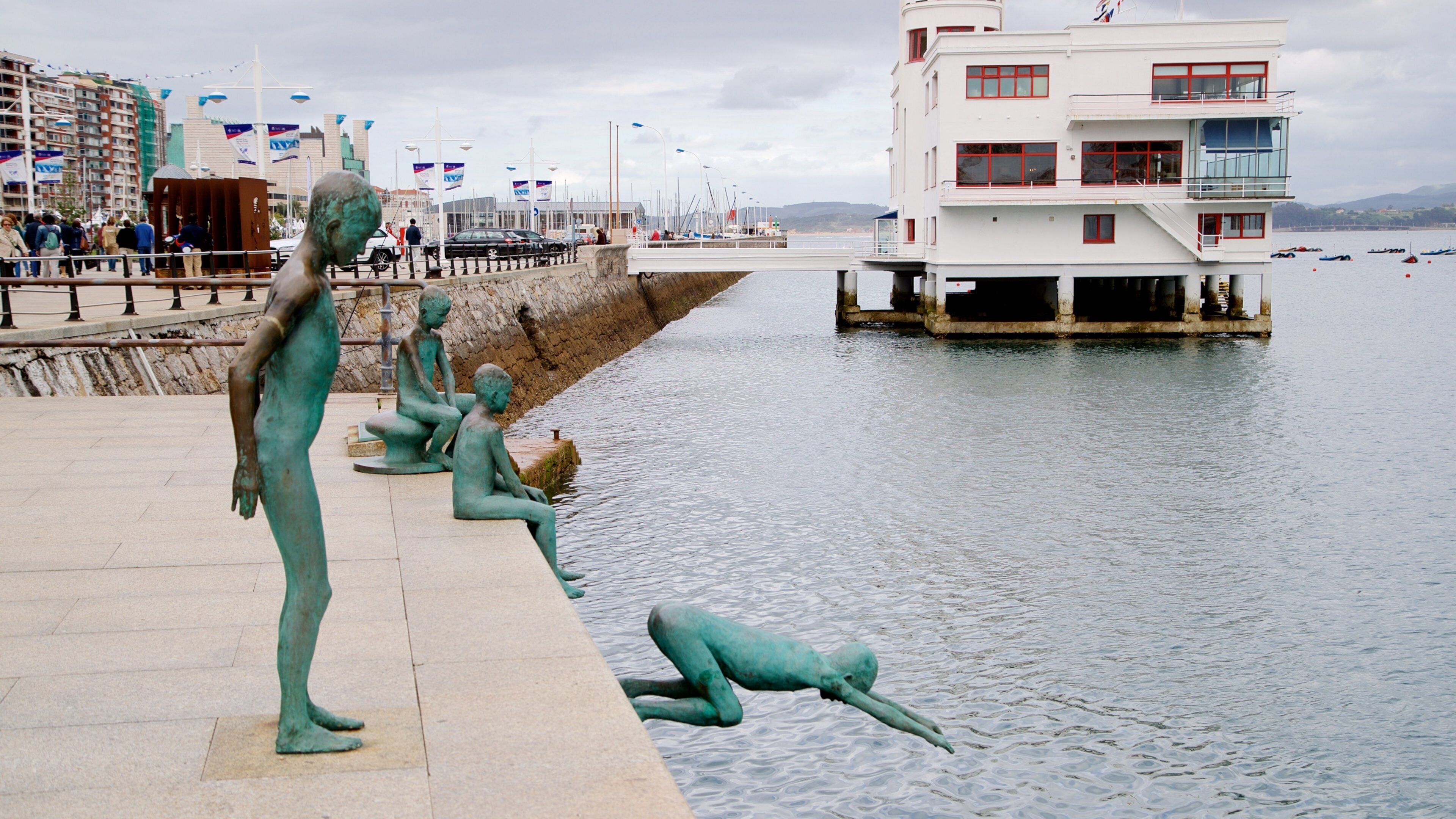 A waterfront promenade with statues of children by the water, a pier with boats, and a white building with red accents.