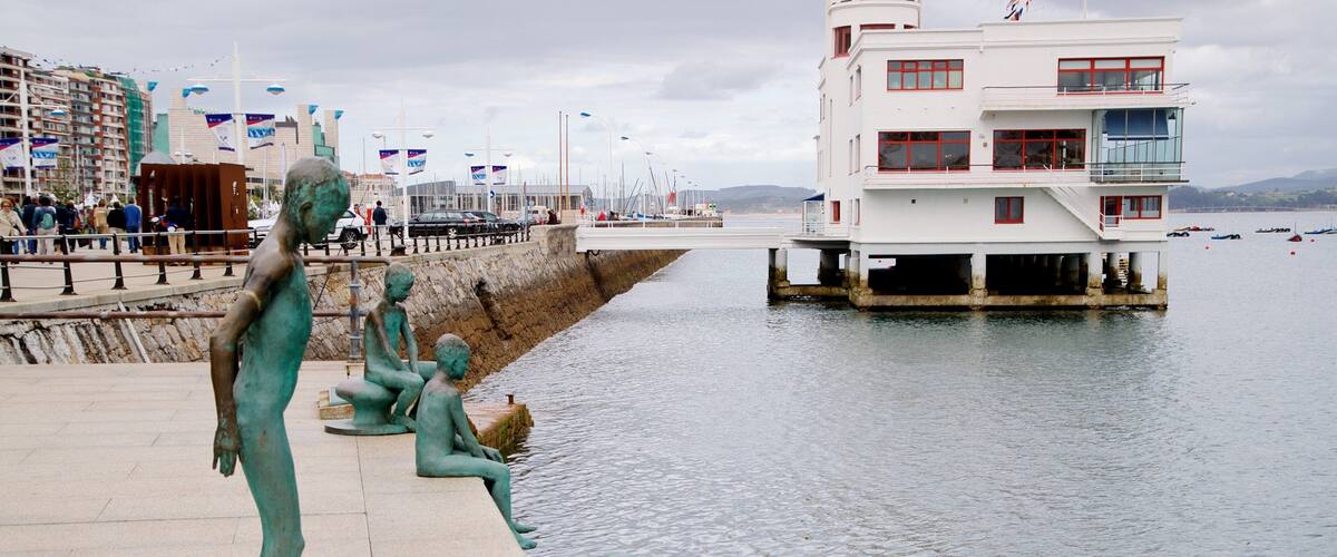 A waterfront promenade with statues of children by the water, a pier with boats, and a white building with red accents.