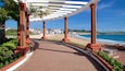 Une promenade dallée, agrémentée de colonnes rouges et surmontée d’un auvent blanc, mène à une plage où l’on aperçoit des bâtiments en arrière-plan.