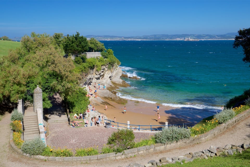 A coastal beach with people sunbathing and swimming, surrounded by greenery and a stone pathway.
