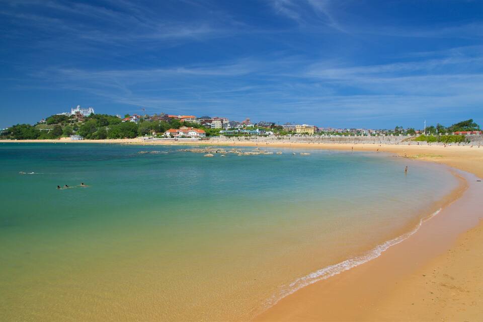 A beach with clear turquoise water, a sandy shore, and buildings in the background.