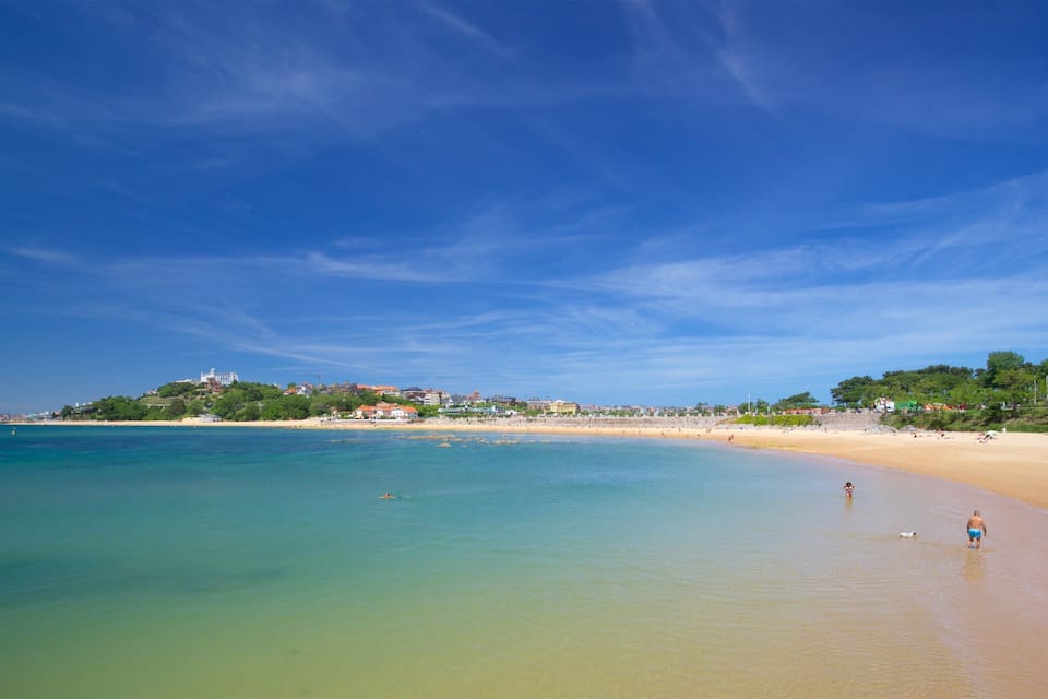 Una playa con aguas turquesas cristalinas, una orilla arenosa y edificios en una colina al fondo.