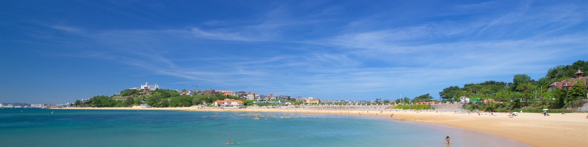 A beach with clear turquoise water, a sandy shore, and buildings on a hill in the background.