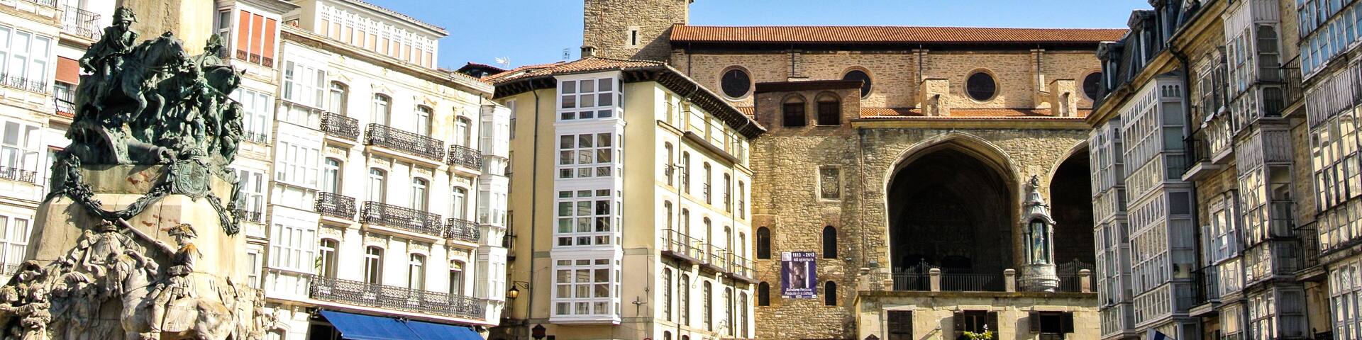 A historic square with a prominent clock tower, a statue of a winged figure, and classical architecture.