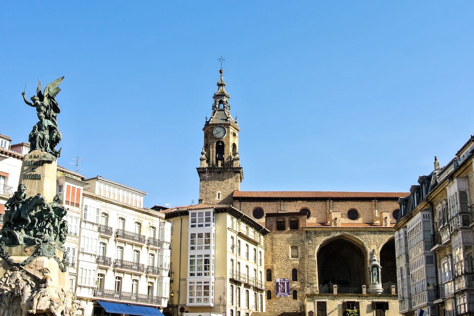Una plaça històrica amb una torre del rellotge destacada, una estàtua d'una figura alada i arquitectura clàssica.