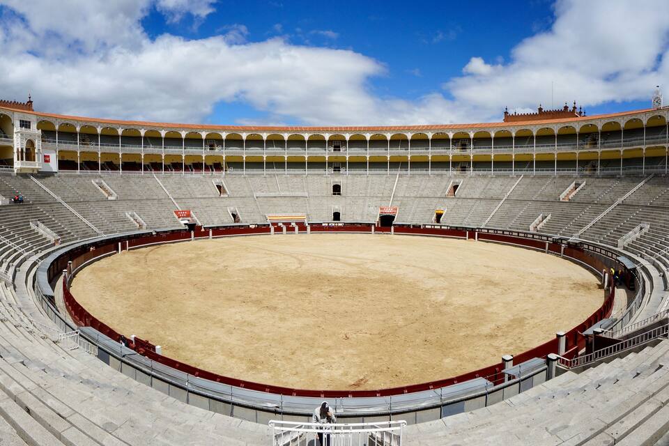 A large, empty bullring with tiered seating and a sandy arena.