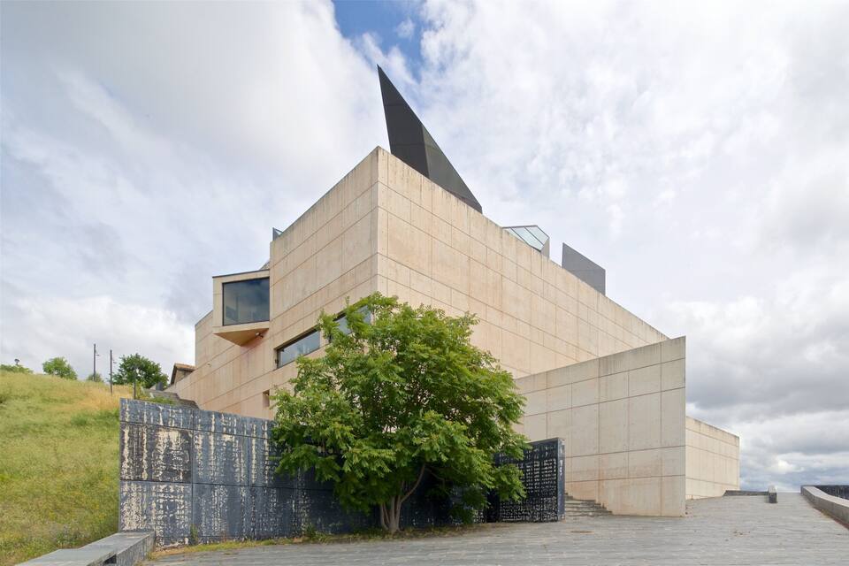 A modern architectural building with a unique triangular design, a tree in front, and a cloudy sky.