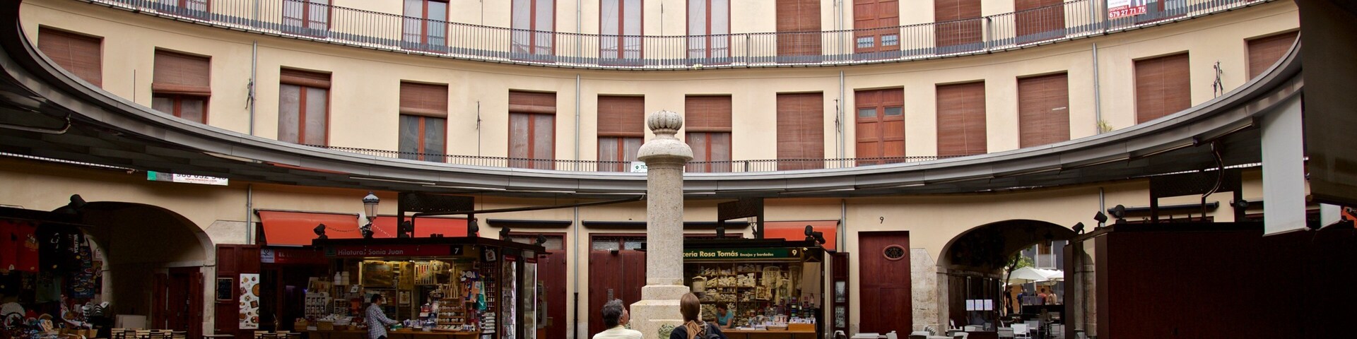 Uma praça circular com uma fonte central, rodeada por edifícios altos com varandas e lojas.