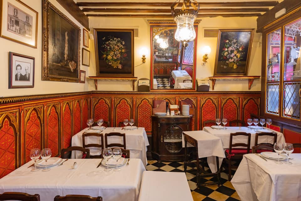 L’intérieur d’un restaurant, avec des boiseries murales richement décorées de motifs rouges et dorés, des nappes blanches et des verres à vin disposés sur les tables.