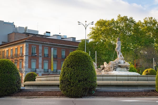 Uma fonte com uma estátua, rodeada por arbustos aparados, em frente a um edifício com uma placa amarela.