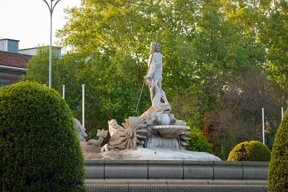 Une statue classique, placée sur une fontaine, entourée de verdure et de bâtiments.