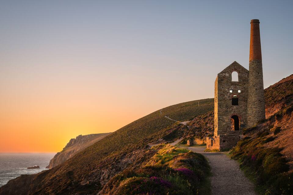 Un paisaje costero con una estructura de piedra en ruinas que presenta una chimenea alta, un camino sinuoso y una puesta de sol sobre el mar.