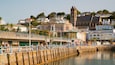 Un pueblo costero con una torre de iglesia destacada, un muelle con gente caminando y diversos edificios, entre ellos una cafetería y un restaurante de mariscos.