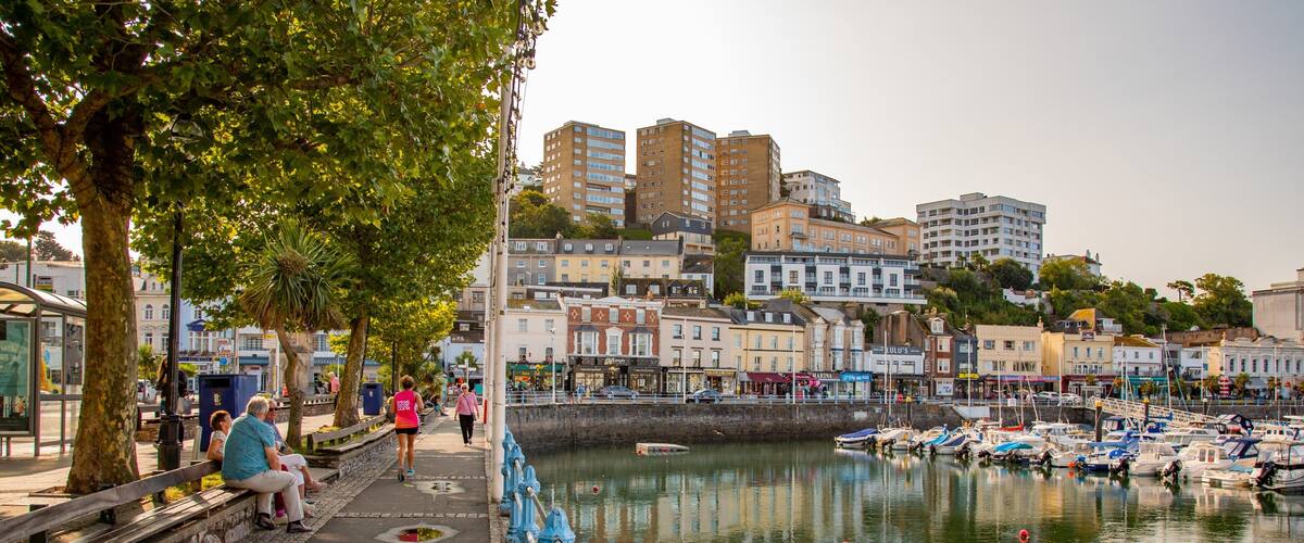A waterfront promenade with people walking, a row of colorful buildings, and boats docked in the water.