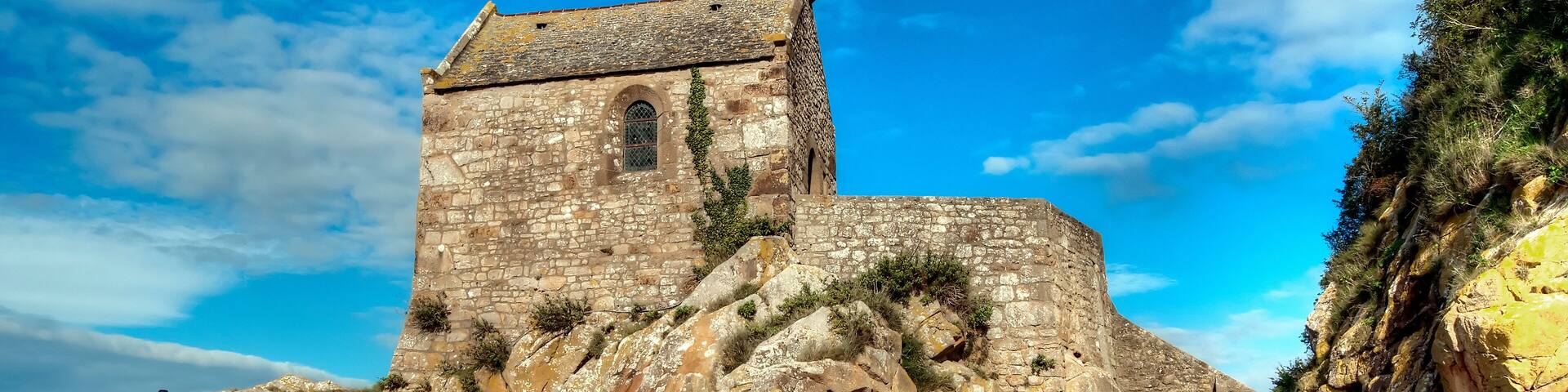 A historic stone church on a rocky cliff overlooking a beach.