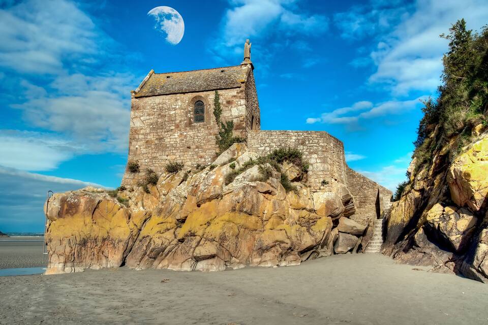 A historic stone church on a rocky cliff overlooking a beach.