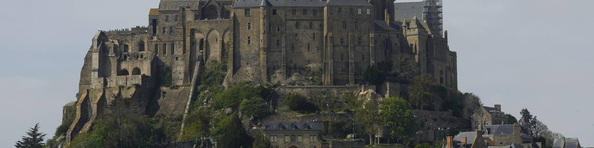 A historic castle with a tall spire, surrounded by lush greenery and smaller buildings.