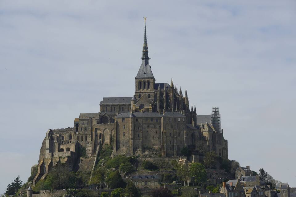 A historic castle with a tall spire, surrounded by lush greenery and smaller buildings.