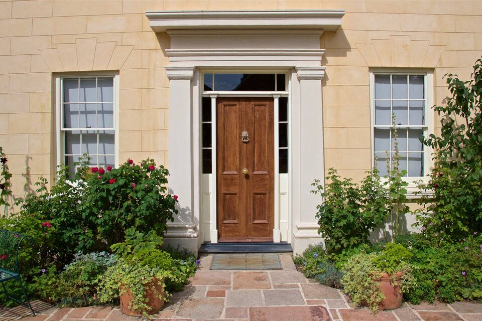 A beige building with a wooden double door, two windows, and a stone pathway leading to the entrance.