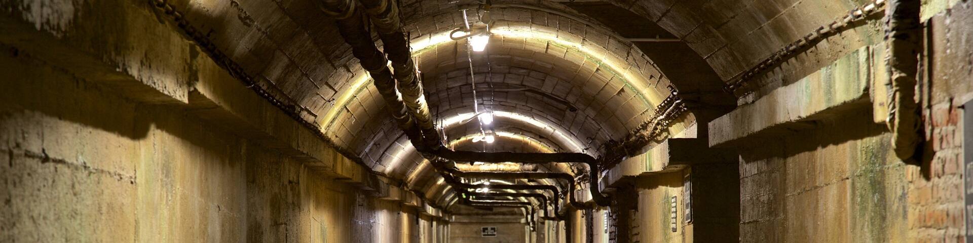 A long, arched tunnel with concrete walls and a metal ceiling, featuring exposed pipes and a light fixture.