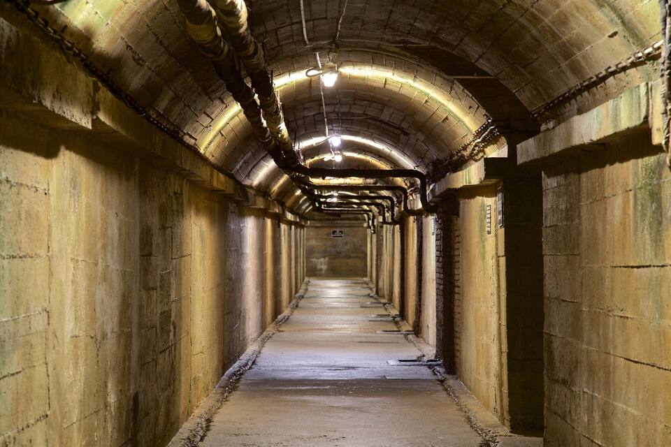 En lang, bueformet tunnel med betonvægge og et metalloft, hvor rør og en lysarmatur er synlige.