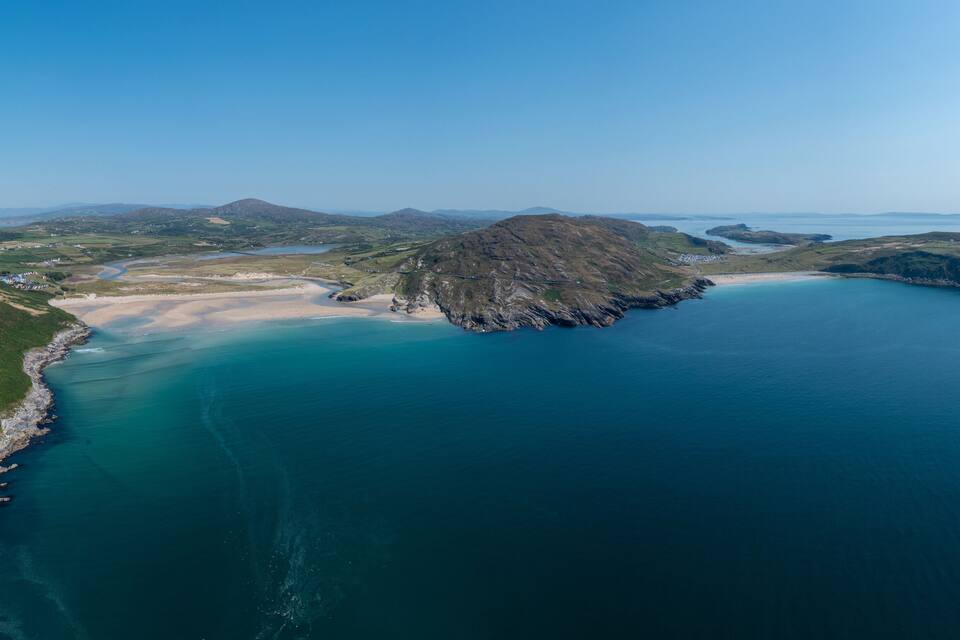 An aerial view of a coastal landscape with a sandy beach, a rocky outcrop, and a clear blue sea.