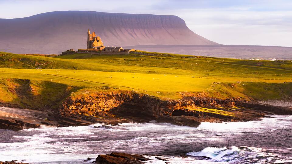 A coastal landscape with a castle on a hill, rocky shoreline, and a large cliff in the background.