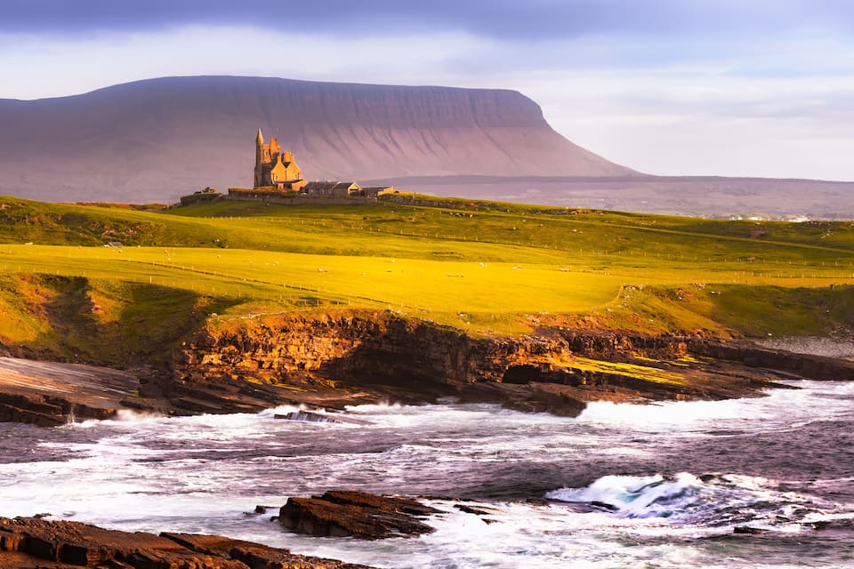 A coastal landscape with a castle on a hill, rocky shoreline, and a large cliff in the background.