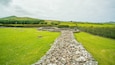A stone wall leading through a green field with a hill in the background.