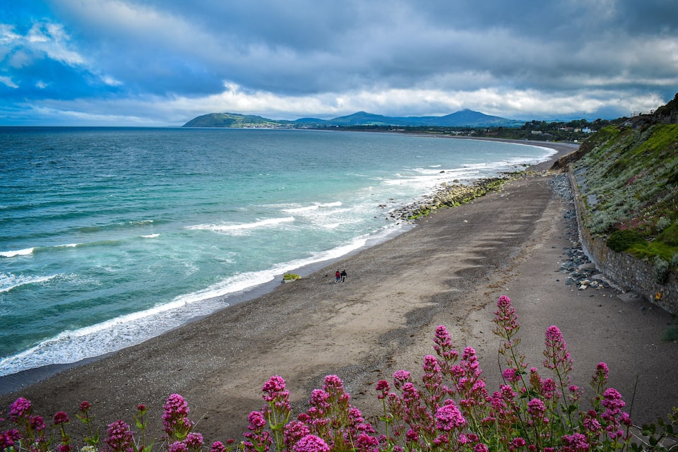 A coastal beach with waves, a person walking, and mountains in the distance.