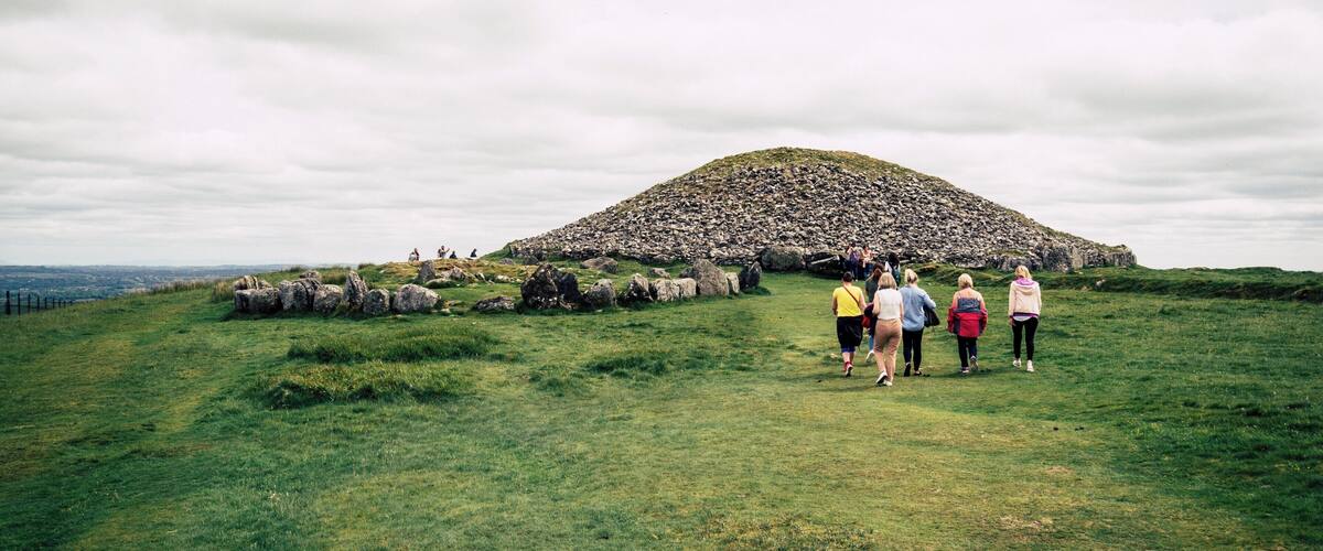 A grassy field with a large mound of stones and a few people walking towards it.