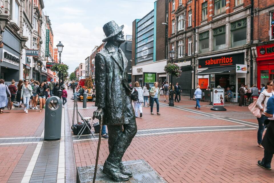 Uma rua urbana movimentada com uma estátua proeminente de um homem usando chapéu e casaco.