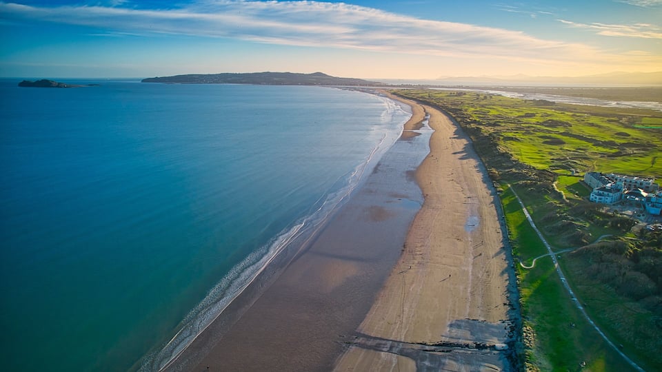 A coastal landscape with a sandy beach, a clear blue sea, and green fields.