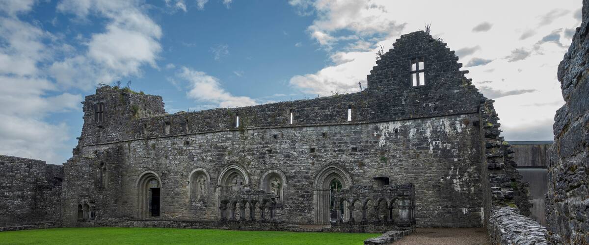 Ruins of an old stone building with arched windows at Abbey Cong, Ireland, surrounded by a lawn under a partly cloudy sky