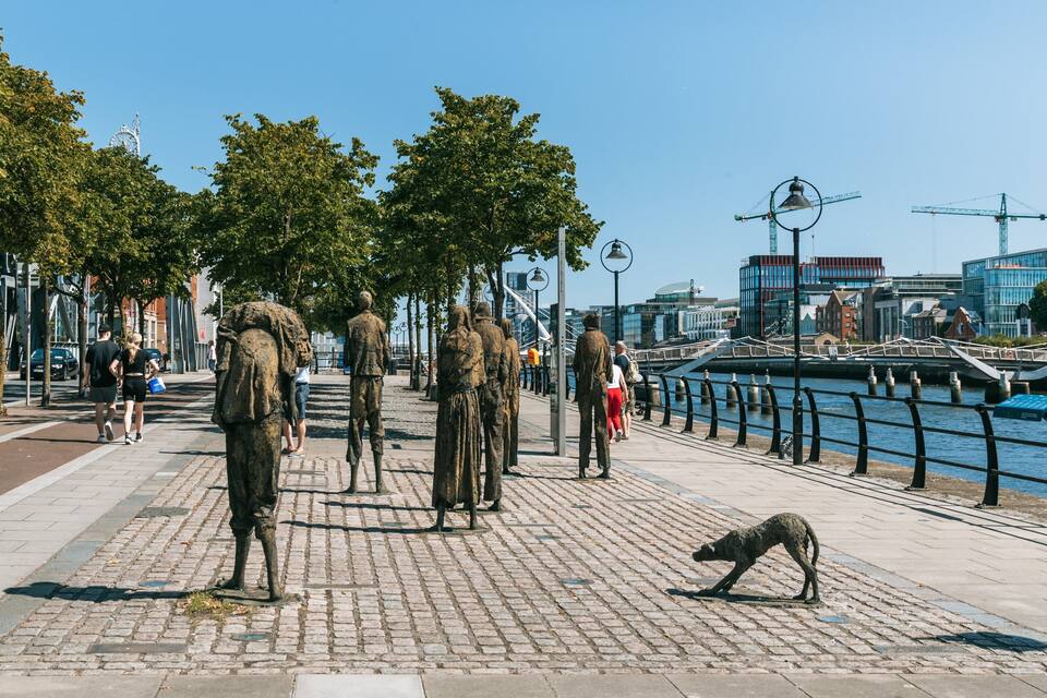 A riverside walkway with statues, a dog sculpture, and modern buildings in the background.