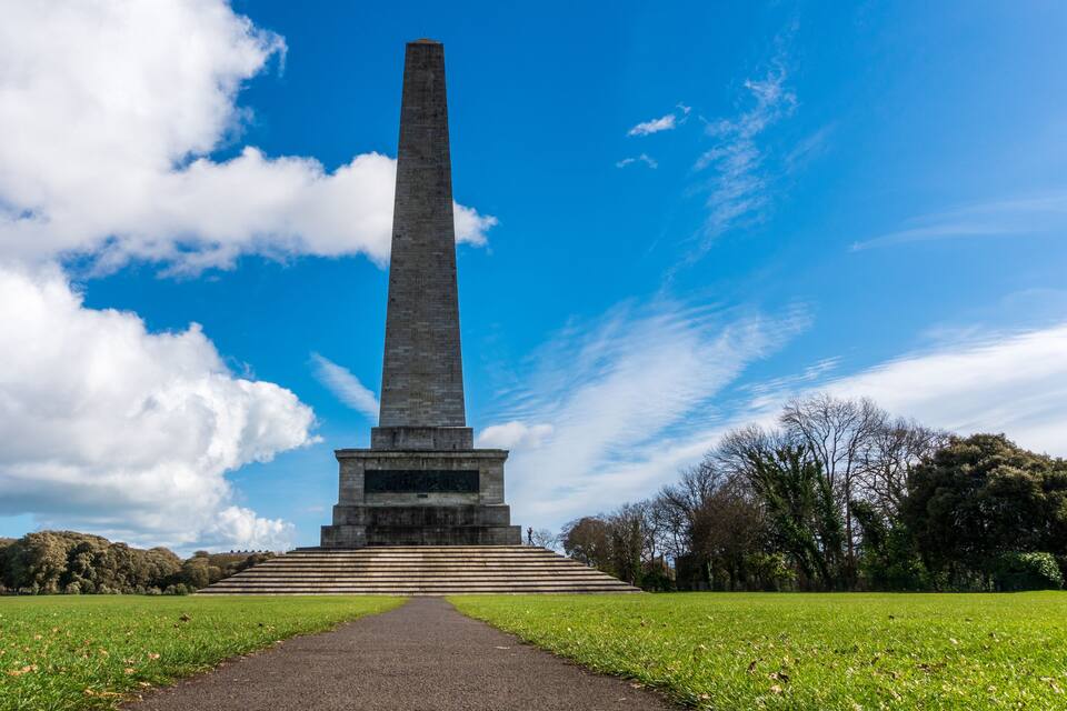 A tall, narrow monument with a broad base, surrounded by a grassy field and trees under a blue sky with clouds.