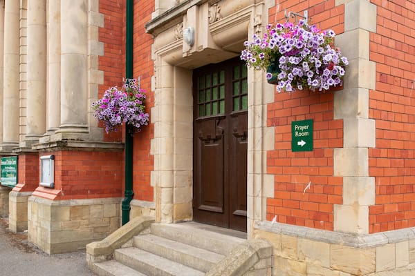 A building with a brick facade, a green sign indicating a prayer room, and flower baskets on the wall.