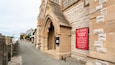 A narrow street with a stone building featuring a large arched entrance. A red sign on the building's wall lists services and contact information for St Paul's Church.