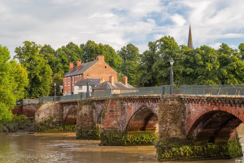 A stone bridge over a river, a brick building, and a church spire in the background.