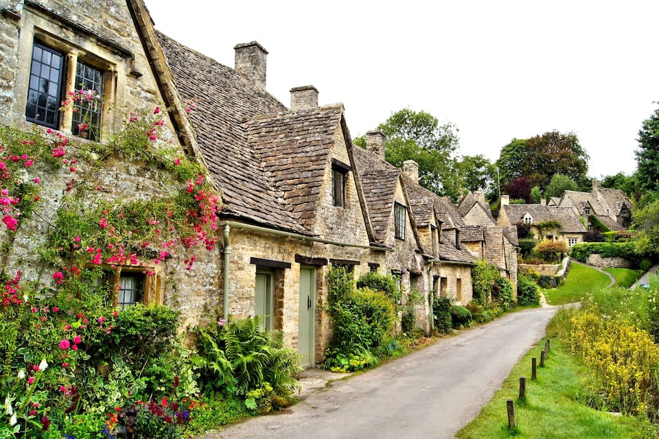 A row of stone cottages with steeply pitched roofs, a narrow road, and lush greenery.