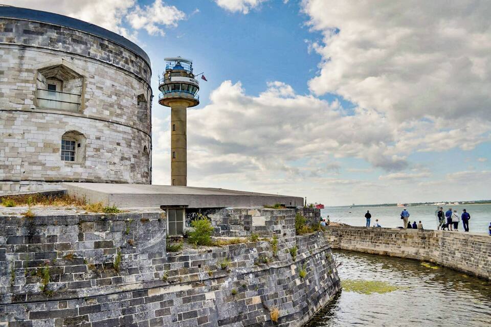 A historic stone building with a lighthouse, a stone wall, and a body of water with people in the background.