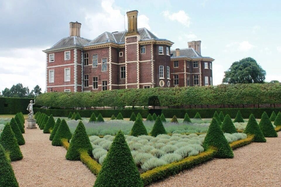 A large brick building with a symmetrical facade and a well-manicured garden with geometrically shaped hedges and a gravel path.