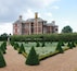 A large brick building with a symmetrical facade and a well-manicured garden with geometrically shaped hedges and a gravel path.