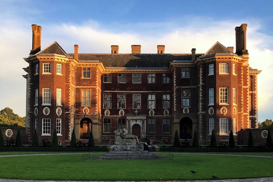 A large, historic brick building with multiple chimneys, a central entrance, and a statue in the foreground.