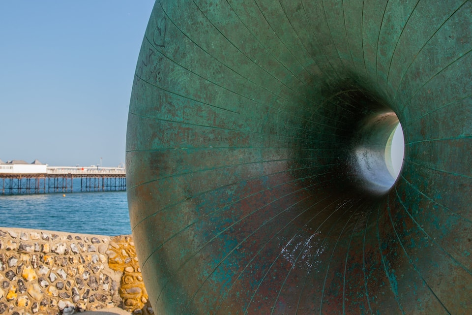 A large greenish metal structure with inscriptions, a stone wall, and a pier extending into the water.