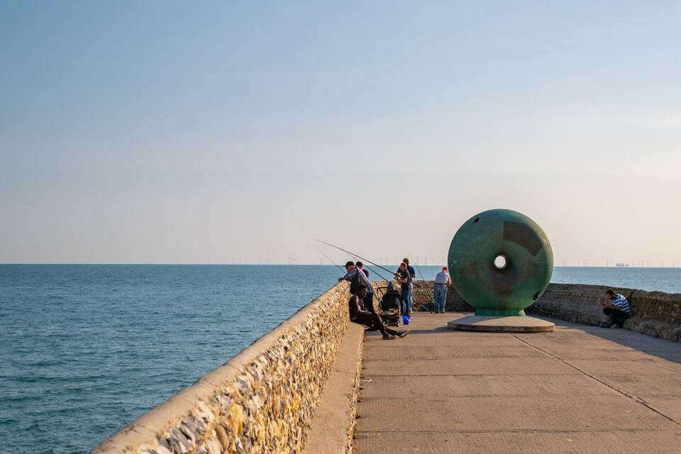 Eine Pier mit Menschen beim Angeln, einer großen grünen Skulptur und ruhigem Meer.