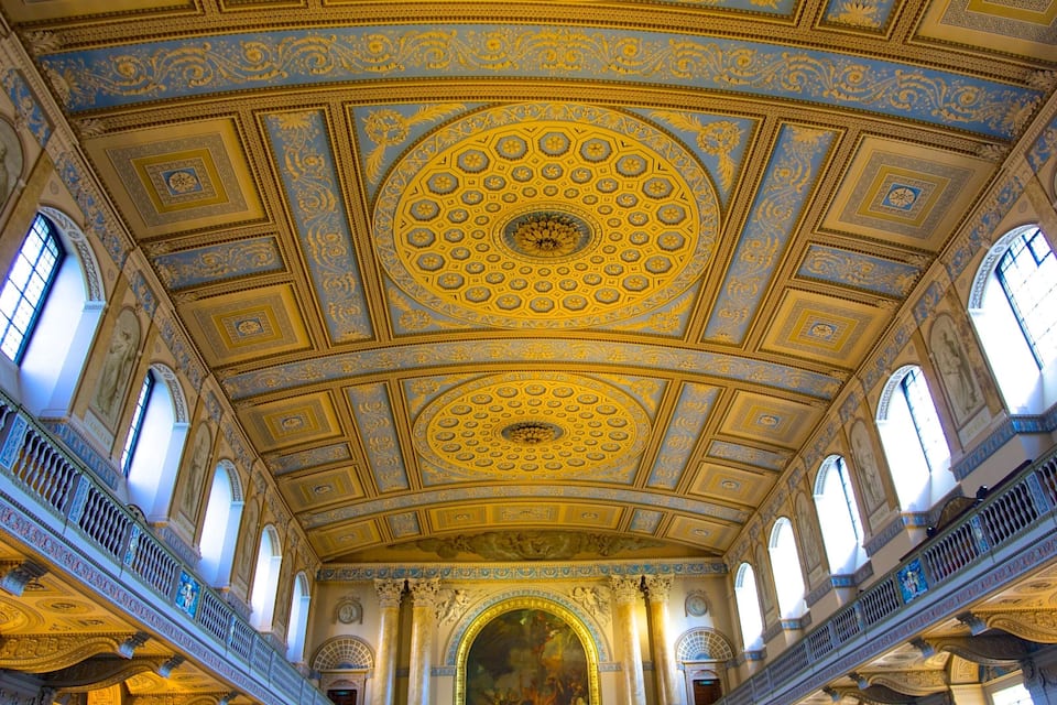 Um salão imponente com teto ricamente decorado em azul e dourado, grandes janelas e um mural central.