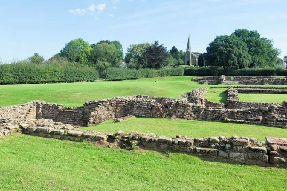 Ancient stone ruins in a grassy field with a church and trees in the background.
