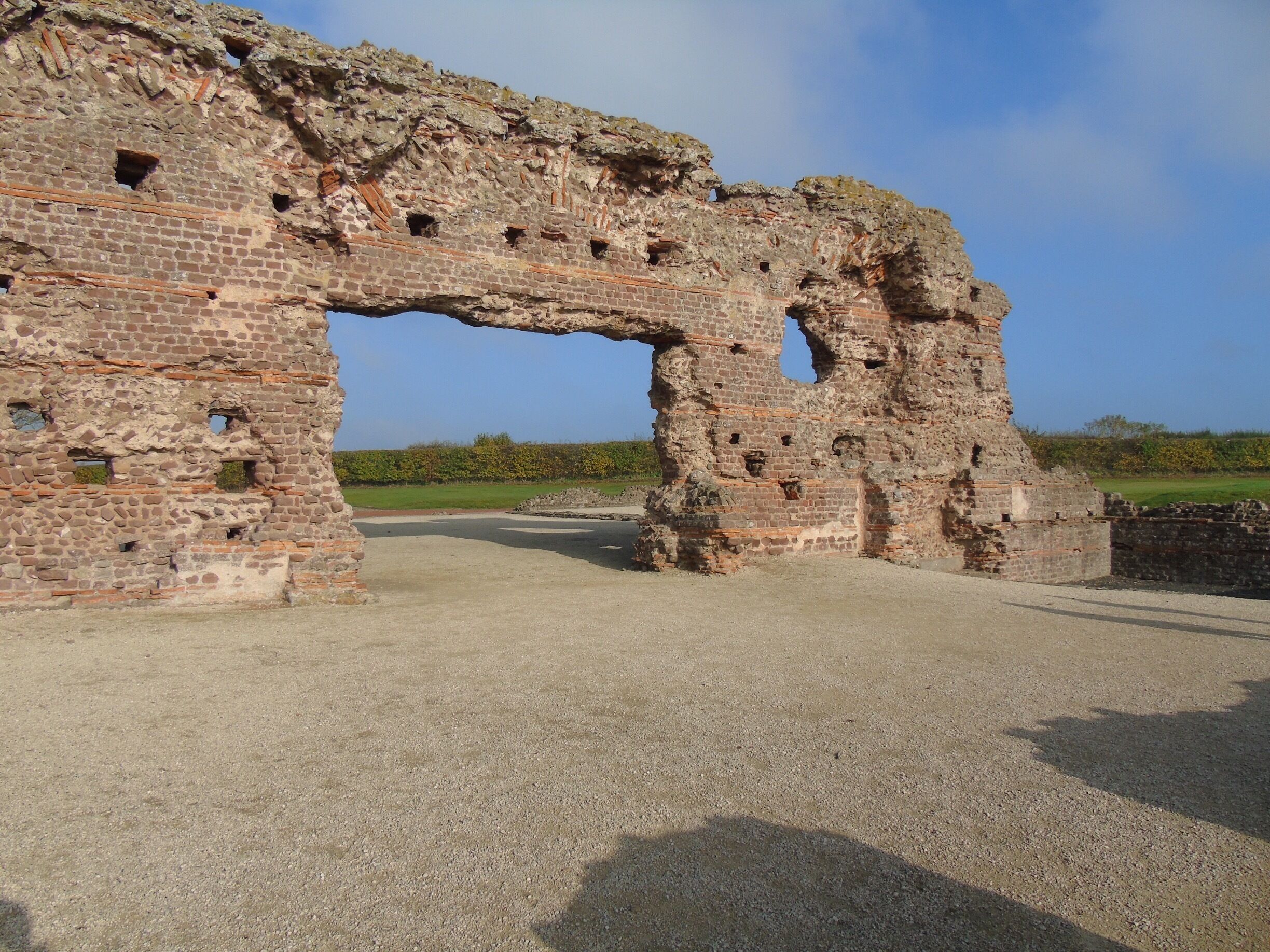 A large, ancient stone archway with visible wear and damage, surrounded by a gravel path and a grassy field.