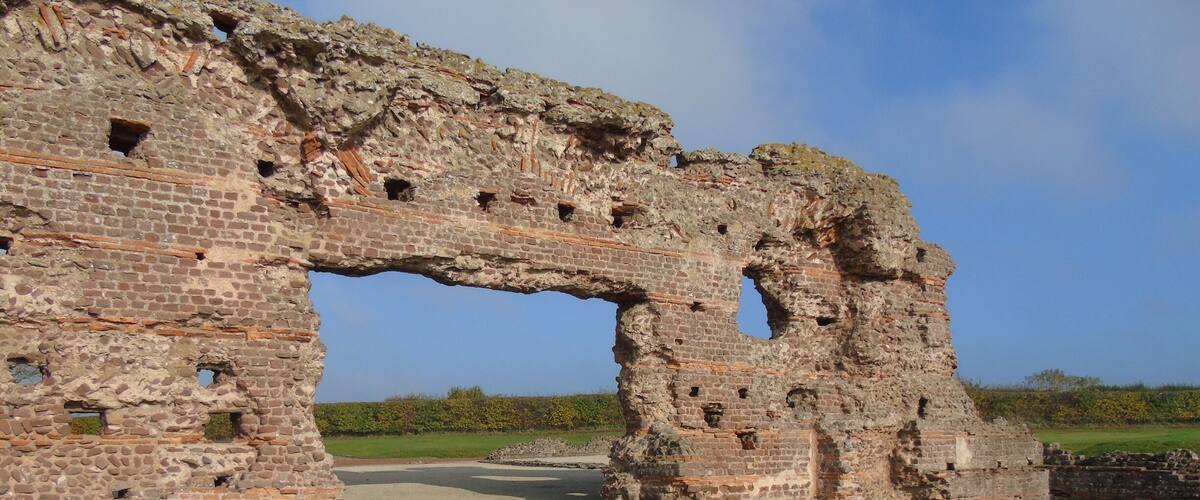A large, ancient stone archway with visible wear and damage, surrounded by a gravel path and a grassy field.
